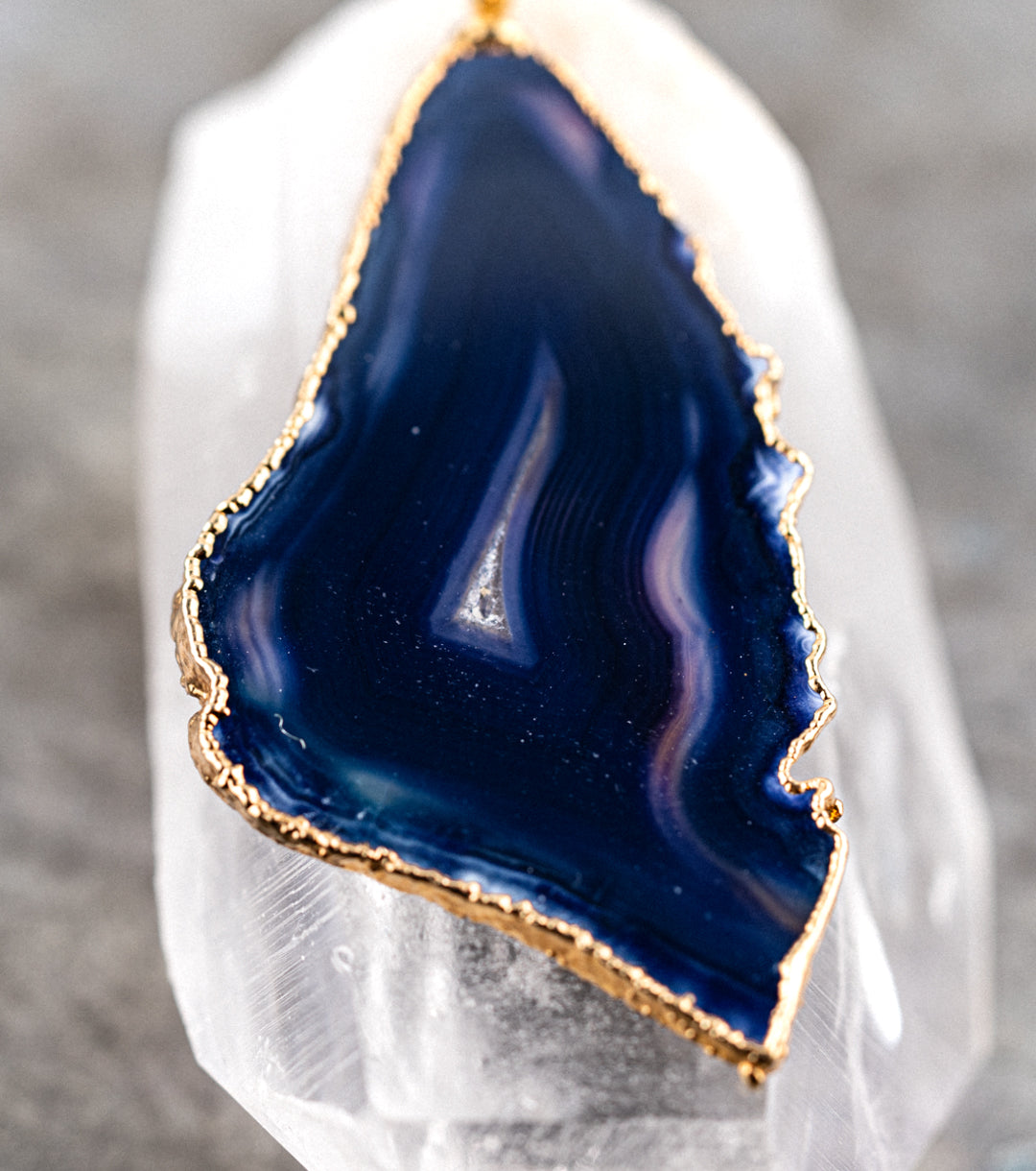 Blue agate pendant with 14K gold wrap and 1.5MM black leather cord necklace on a reflective quartz pedestal against a gray metal background