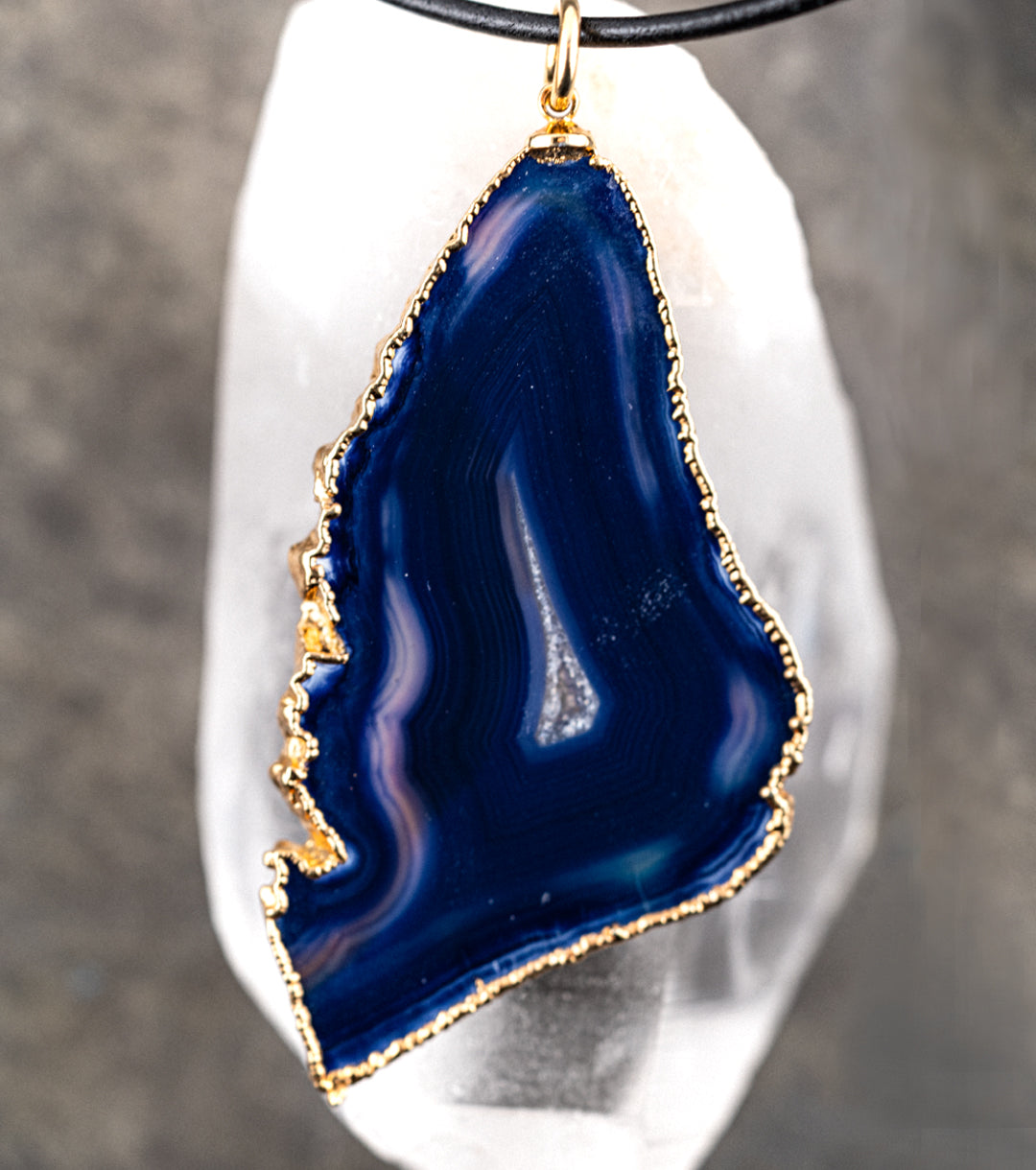 Blue agate pendant with 14K gold wrap and 1.5MM black leather cord necklace on a reflective quartz pedestal against a gray metal background